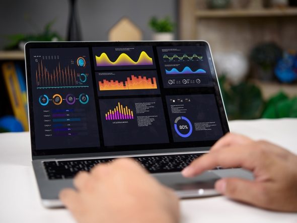 Close-up of a laptop whose screen displays a dashboard with multiple colorful graphs, charts and statistical metrics as hands tap on it. The image visualizes business intelligence (BI), data analysis, real-time reporting, business performance and performance monitoring.
