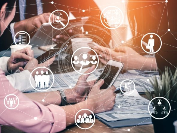 A team of business people sits around a table while a digital network of glowing icons for communication, employees and organization (e.g. teamwork, HR, documents) lies above them. The image visualizes teamwork, business communication, digital collaboration, networking and business relationships.