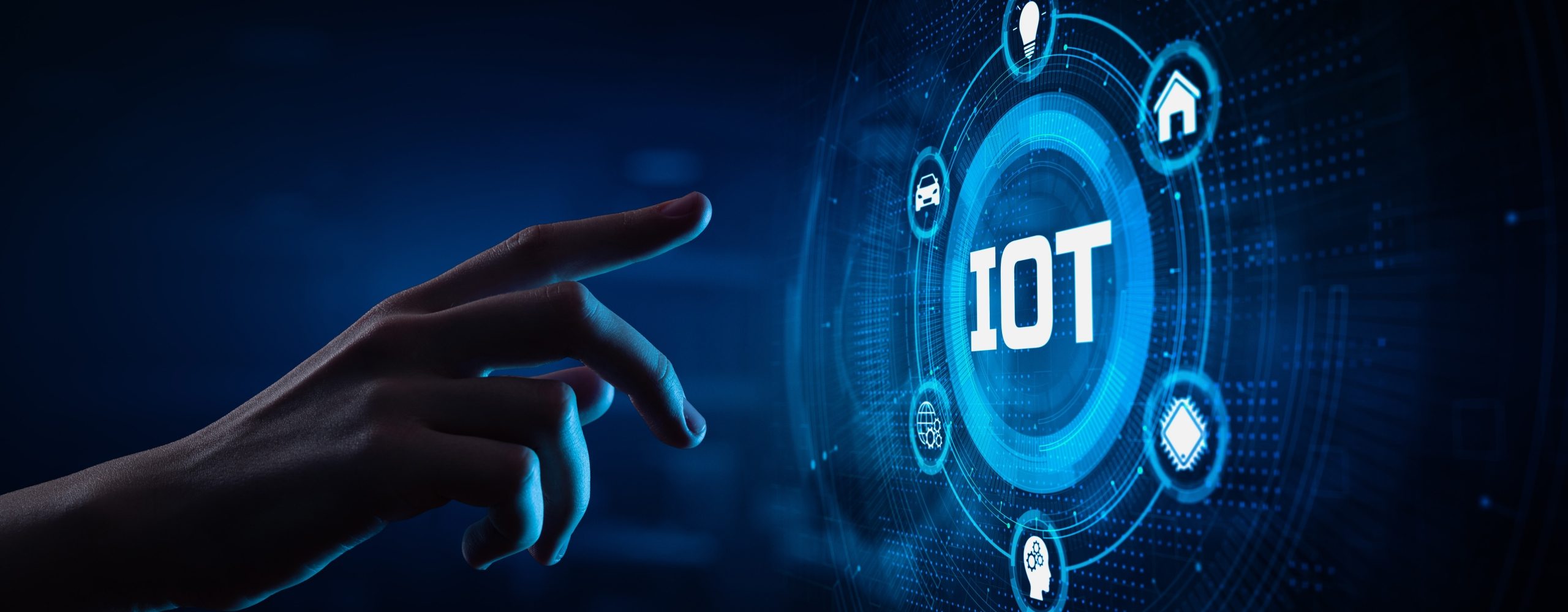Eine menschliche Hand berührt ein futuristisches, leuchtend blaues Digital-Display mit dem zentralen Schriftzug IOT (Internet of Things). Um das IOT-Symbol sind Icons angeordnet, die vernetzte Geräte wie Auto, Haus, Sensor und Kopf (Künstliche Intelligenz) symbolisieren. Das Bild veranschaulicht die digitale Vernetzung im Zeitalter des Internet der Dinge.
