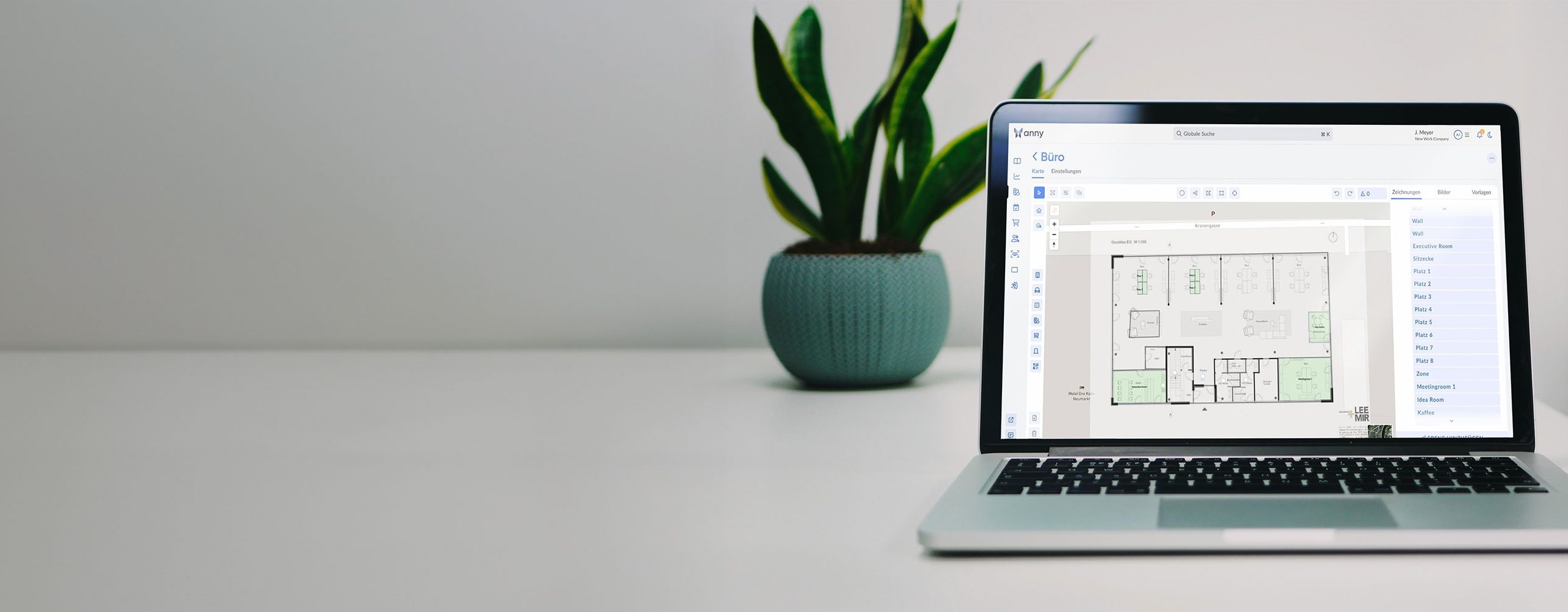 Laptop auf einem weißen Schreibtisch mit einer Zimmerpflanze im Hintergrund. Auf dem Bildschirm ist die anny Buchungssoftware zu sehen, die einen Büro-Grundriss für Desk Sharing oder Raummanagement darstellt.