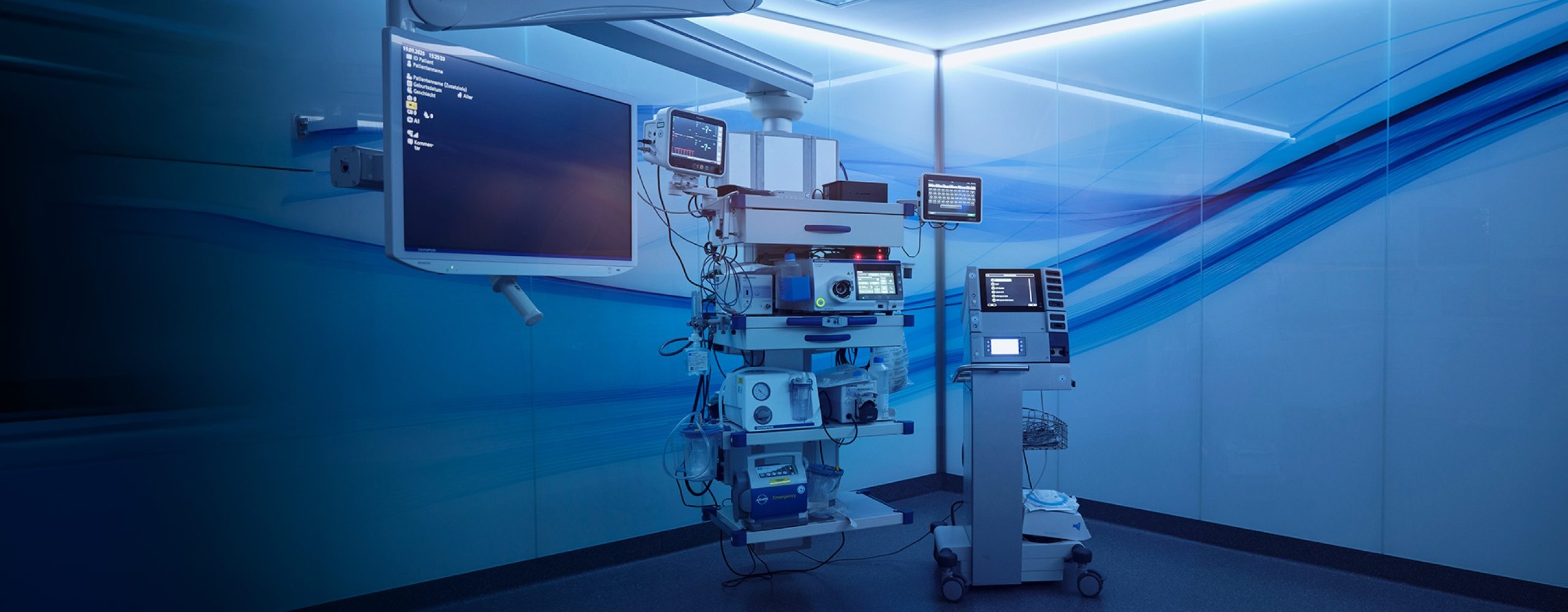 Ein moderner, futuristisch gestalteter Operationssaal oder Behandlungsraum mit medizinischen Geräten auf einem Rollwagen, Monitoren und blau leuchtenden Wandpanelen mit Wellenmuster.
