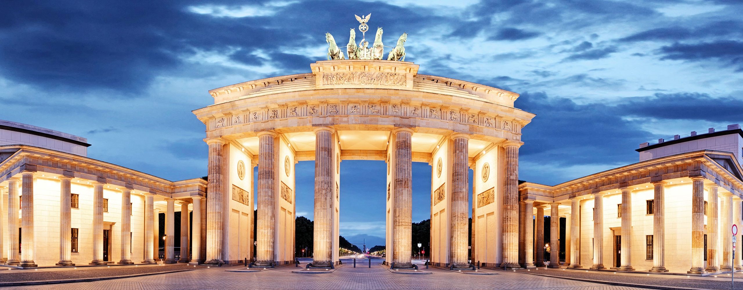 Das Brandenburger Tor in Berlin in der Dämmerung, beleuchtet und von dunklen Wolken umrahmt. Die Nachtansicht betont die imposante Säulenarchitektur und die Quadriga auf der Spitze.