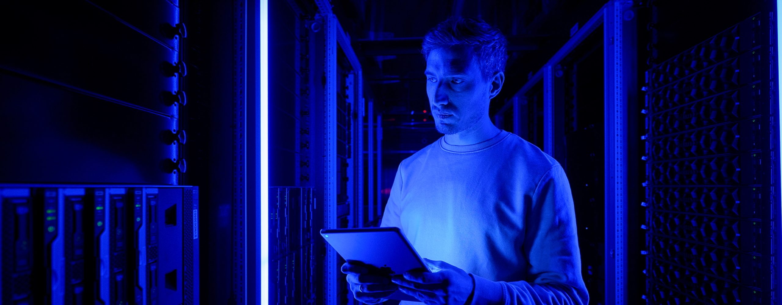A man holding a glowing tablet, looking at server racks in a dark data center with vibrant blue neon lighting.