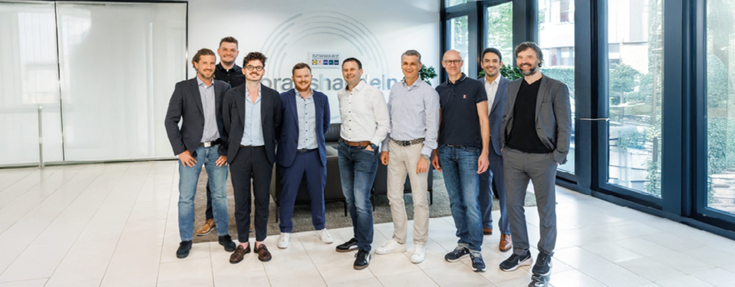 Gruppe von neun professionellen Männern, die in einer modernen Büro-Lobby mit großen Fenstern und einem Schwarz-Blockchain-Logo an der Wand stehen.