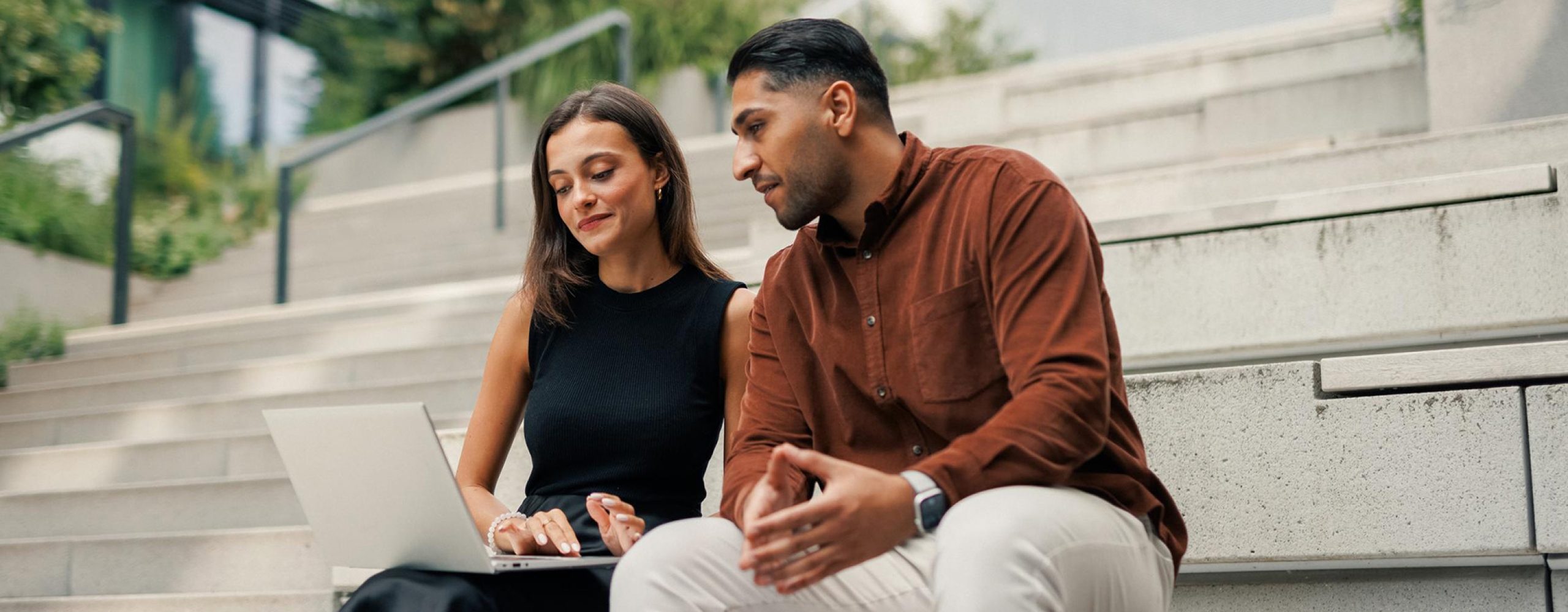 Two colleagues are working together on a laptop, sitting on a stone staircase outdoors.