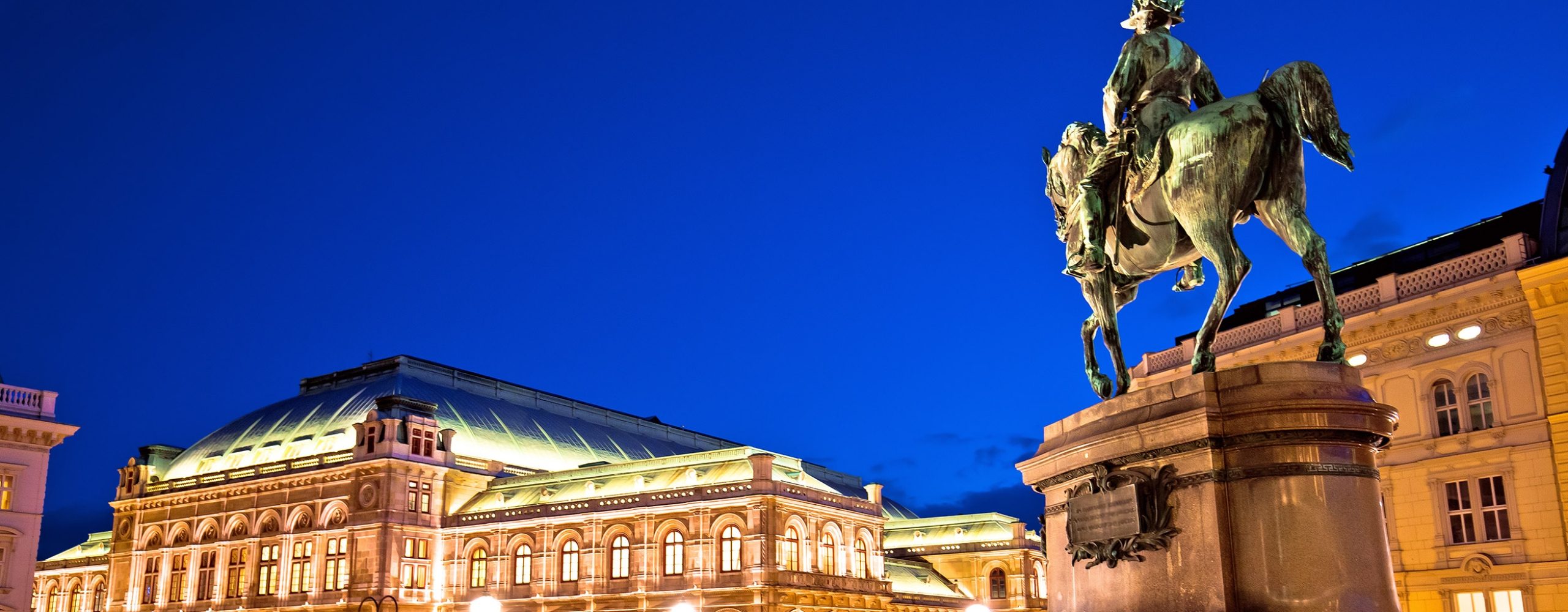 Beleuchtete Wiener Staatsoper bei Nacht mit dem historischen Reiterstandbild auf der Albertinarampe im Vordergrund. Ein imposantes architektonisches Wahrzeichen und eine beliebte Sehenswürdigkeit in Wien vor tiefblauem Abendhimmel.