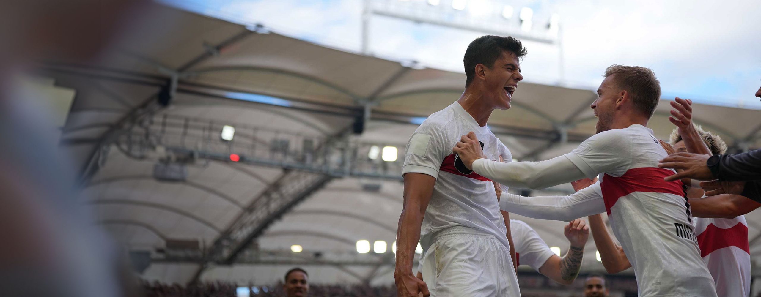 Jubelnde VfB Stuttgart Fußballspieler (in weißen Heimtrikots) nach einem Torerfolg im Stadion. Ein Spieler schreit seine Freude heraus, während er von Teamkollegen umarmt wird.