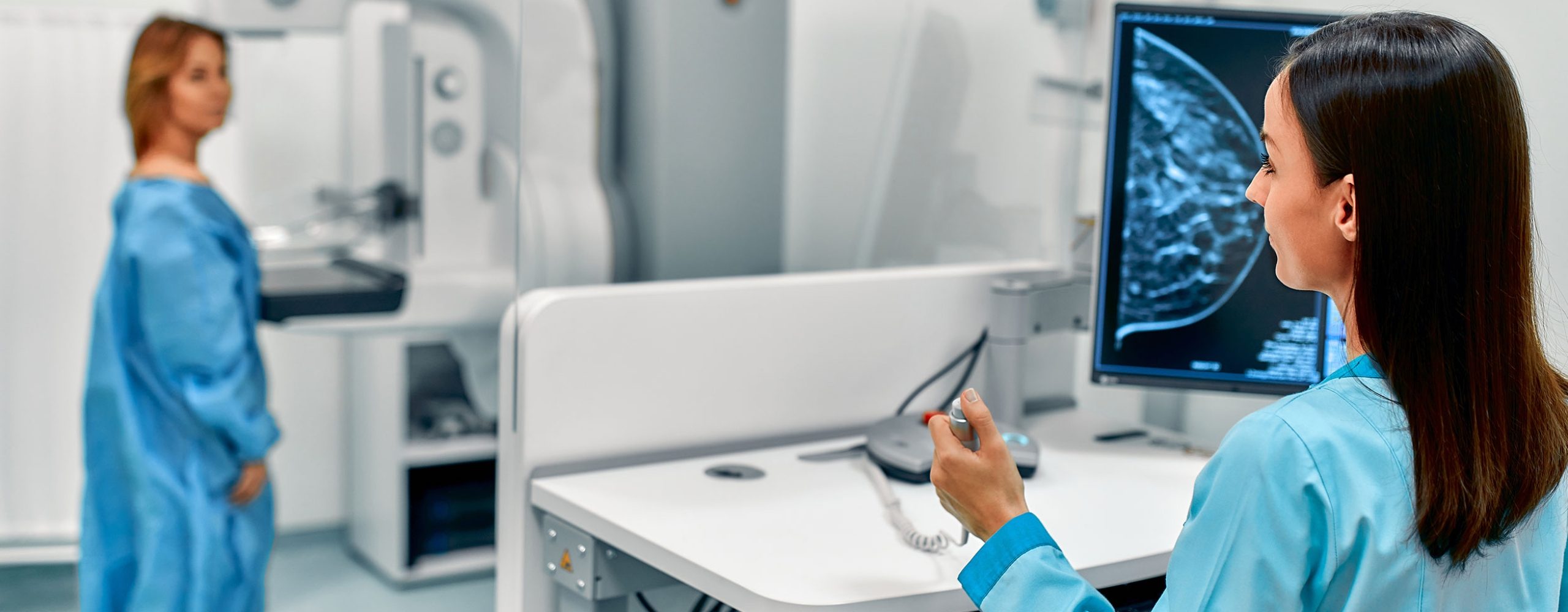Medizinische Fachkraft wertet Mammographie-Aufnahmen am Monitor aus, während eine Patientin am Untersuchungsgerät steht.