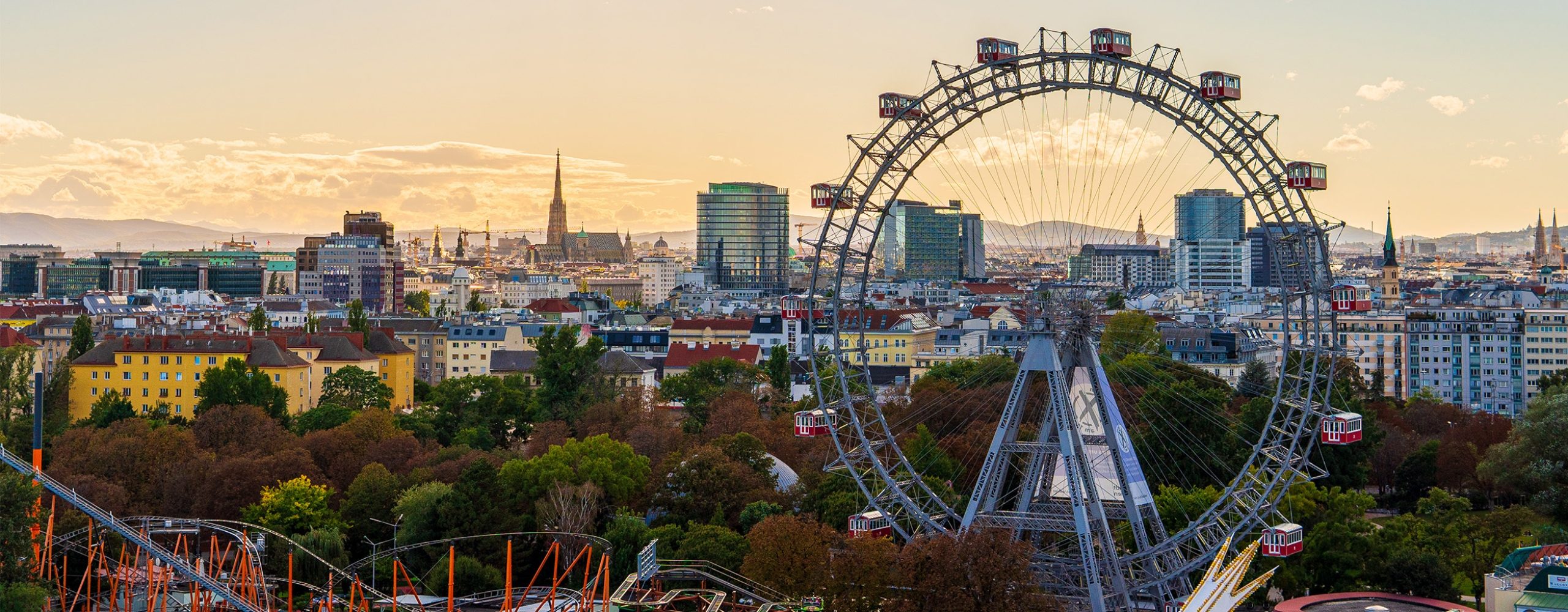 Ein Panoramablick auf die Wiener Skyline während der goldenen Stunde, dominiert vom historischen Wiener Riesenrad im Prater. Im Hintergrund ist der markante Turm des Stephansdoms inmitten der Stadtlandschaft zu sehen, während Sonnenstrahlen durch eine dramatische Wolkendecke am Abendhimmel brechen.