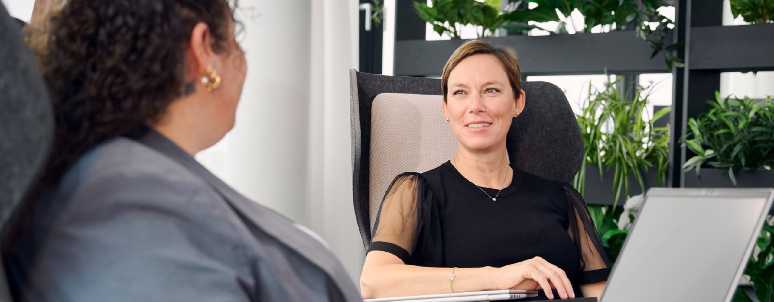 Eine Frau lächelt während einer geschäftlichen Besprechung in einem modernen Büro mit biophilem Design; im Vordergrund eine Kollegin am Laptop als Symbol für moderne IT-Zusammenarbeit.