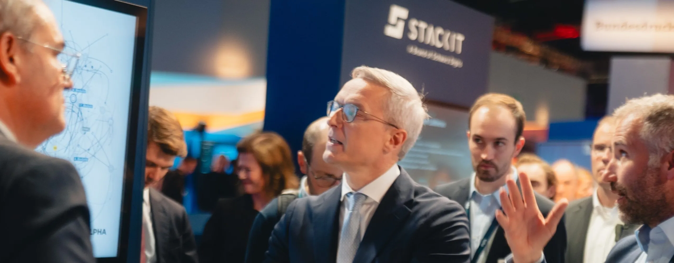 Nahaufnahme von Fachleuten in Anzügen bei einer Präsentation auf einer Technologiemesse. Ein Bildschirm zeigt komplexe Graphen, die ein Netzwerk oder System visualisieren. Die Teilnehmer sind in ein intensives Gespräch vertieft, einer erklärt mit einer Geste. Im Hintergrund sind Firmenlogos wie STACKIT und Bundesdruckerei Gruppe erkennbar, was den geschäftlichen Kontext unterstreicht.
