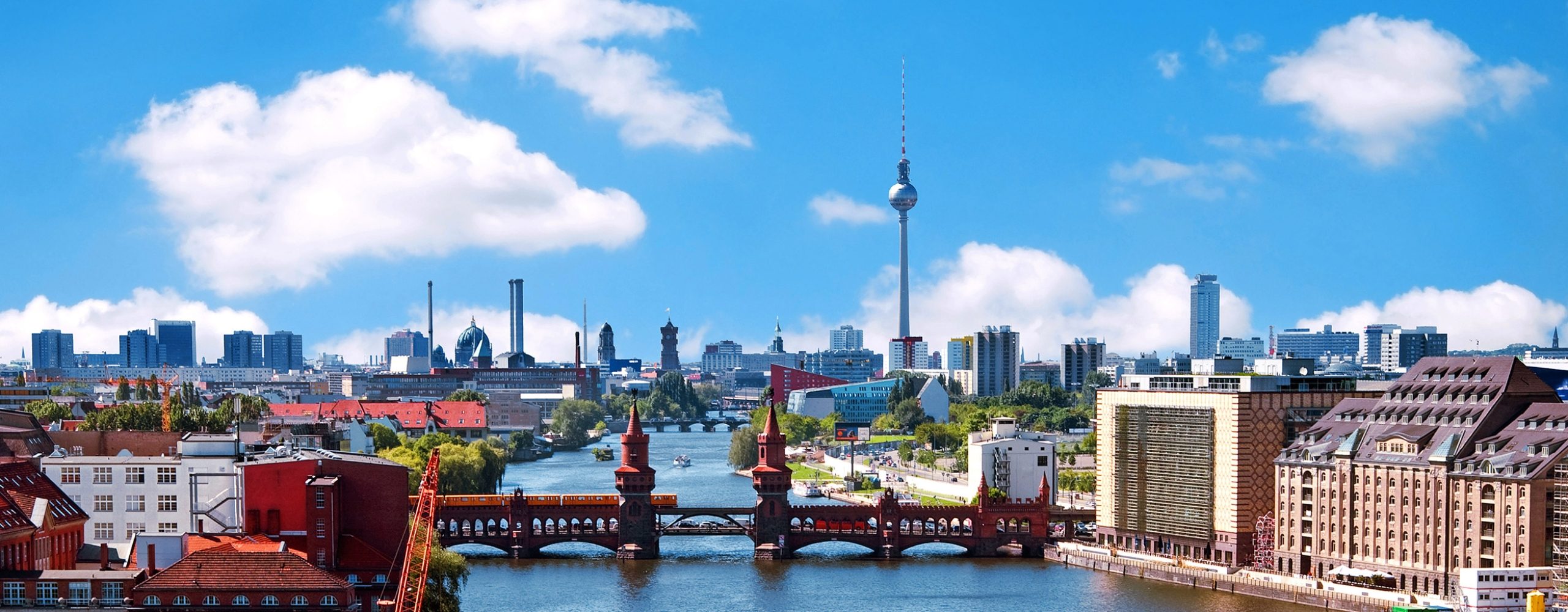 Panorama-Blick auf die Berliner Skyline mit der Oberbaumbrücke über der Spree und dem Berliner Fernsehturm im Hintergrund unter einem blauen Wolkenhimmel. Eine gelbe U-Bahn überquert gerade die Brücke.
