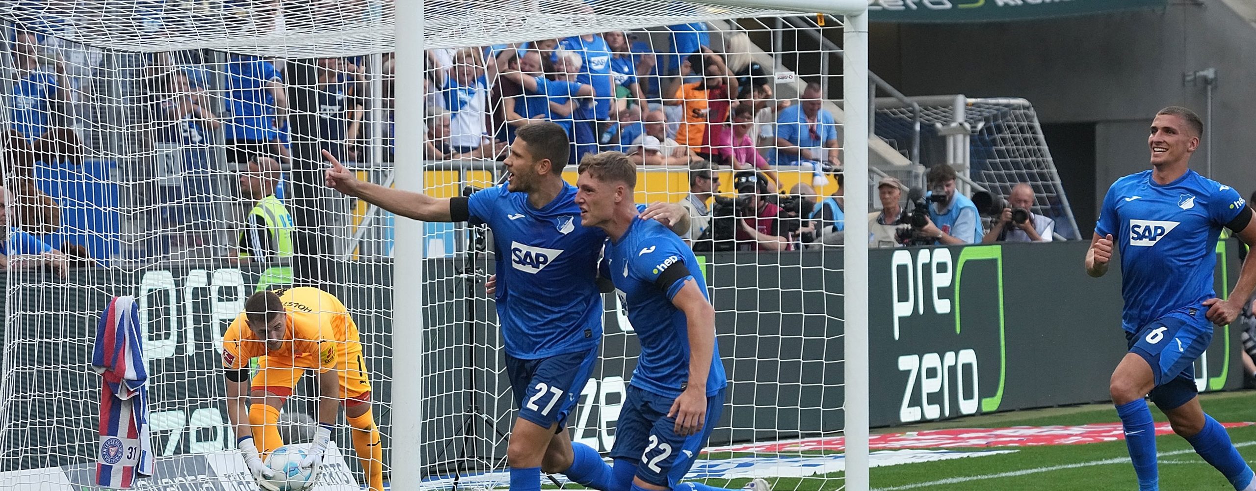 TSG Hoffenheim soccer players (in blue jerseys with numbers 27 and 22) celebrate a goal in front of the net. The opposing goalkeeper is on the ground, and fans cheer in the stands. A "Zero Waste" banner by PreZero is visible in the stadium.