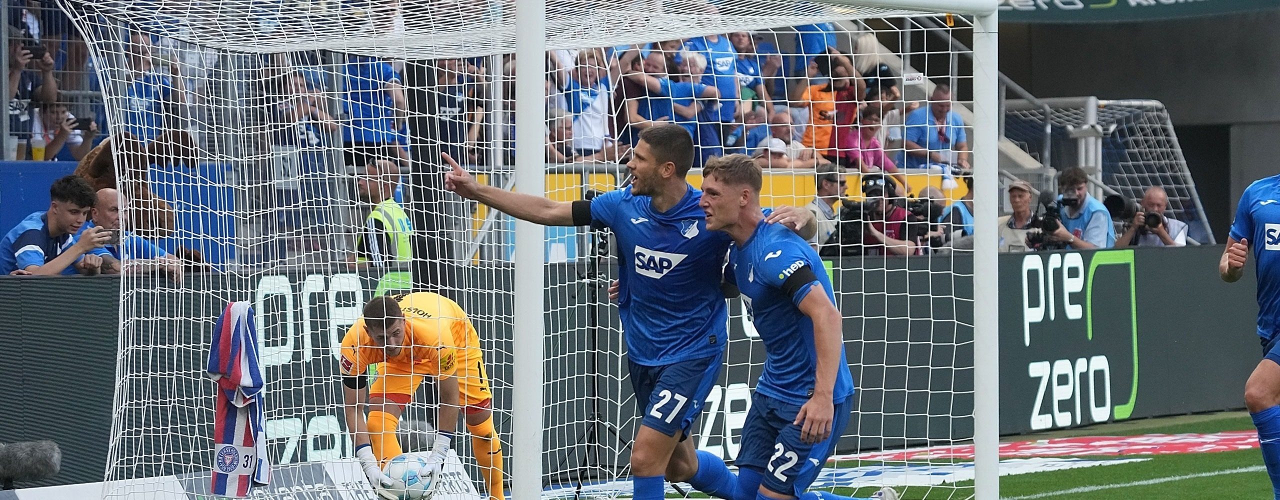Fußballspieler der TSG Hoffenheim (in blauen Trikots mit den Nummern 27 und 22) jubeln vor dem Tor nach einem erzielten Treffer. Der Torwart liegt geschlagen am Boden, im Hintergrund jubeln die Fans. Im Stadion ist das Banner "Zero Waste" von PreZero zu sehen.