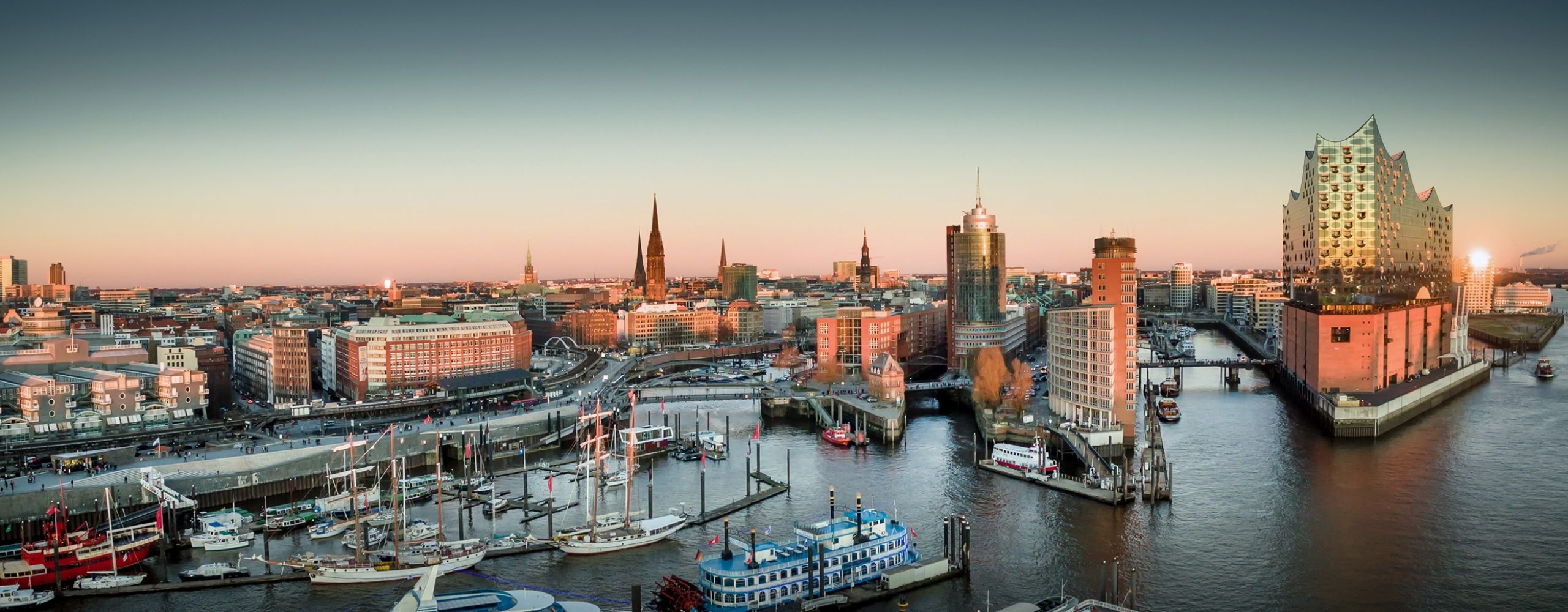 Panoramic view of Hamburg harbor at sunset featuring the Elbphilharmonie building on the right, boats docked in the waterway, and the city skyline in the background.