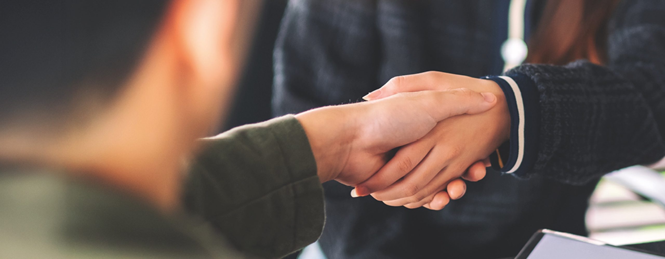 Nahaufnahme von zwei Personen, die sich die Hände schütteln, was eine Geschäftsvereinbarung oder Partnerschaft symbolisiert. Eine Person trägt ein olivgrünes Oberteil, die andere eine Jacke mit gestreiften Manschetten. Der Hintergrund ist verschwommen, mit einem Tisch oder Schreibtisch, der auf ein Büro- oder Besprechungsraum-Setting hindeutet.