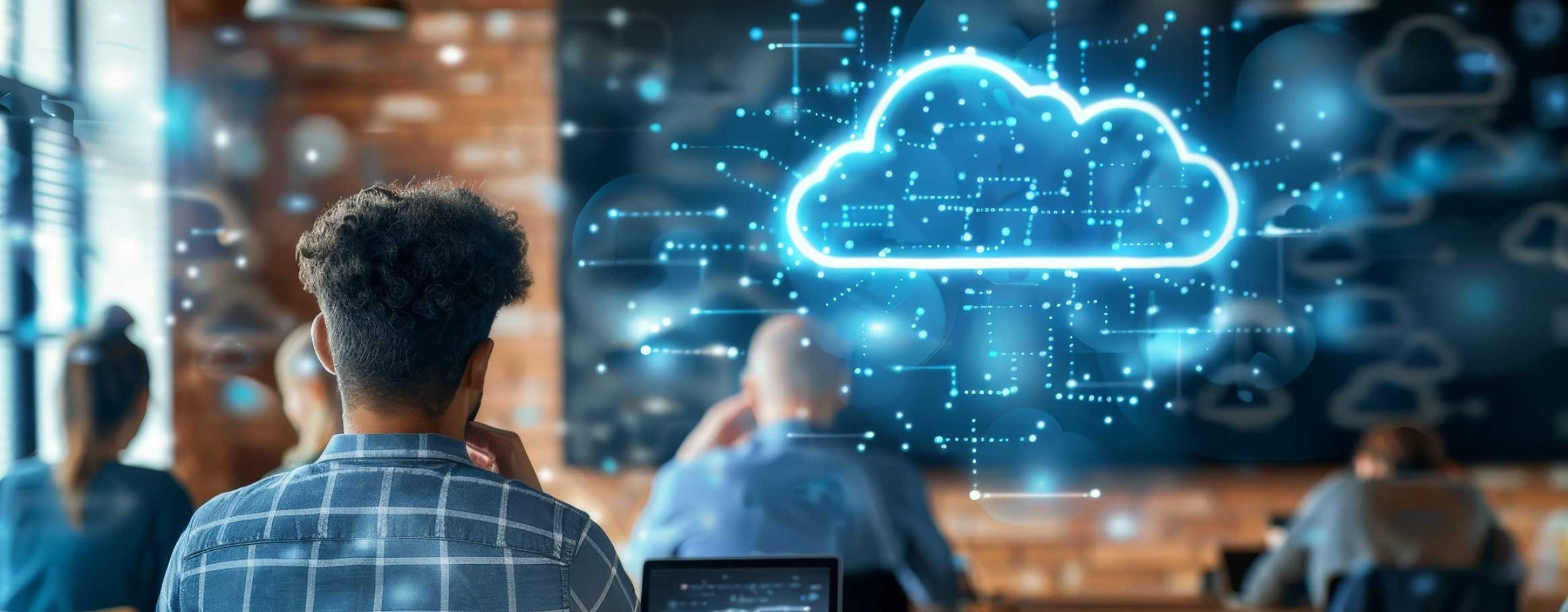 Back view of a man with curly hair sitting in front of his laptop in a modern, rustic office or coworking space. On the screen or as an augmented reality overlay, a bright blue, digitized cloud (cloud symbol) can be seen surrounded by a network of lines and dots. In the blurred background, other people are sitting at long wooden tables, suggesting a training session, webinar or technology presentation centered around cloud computing.