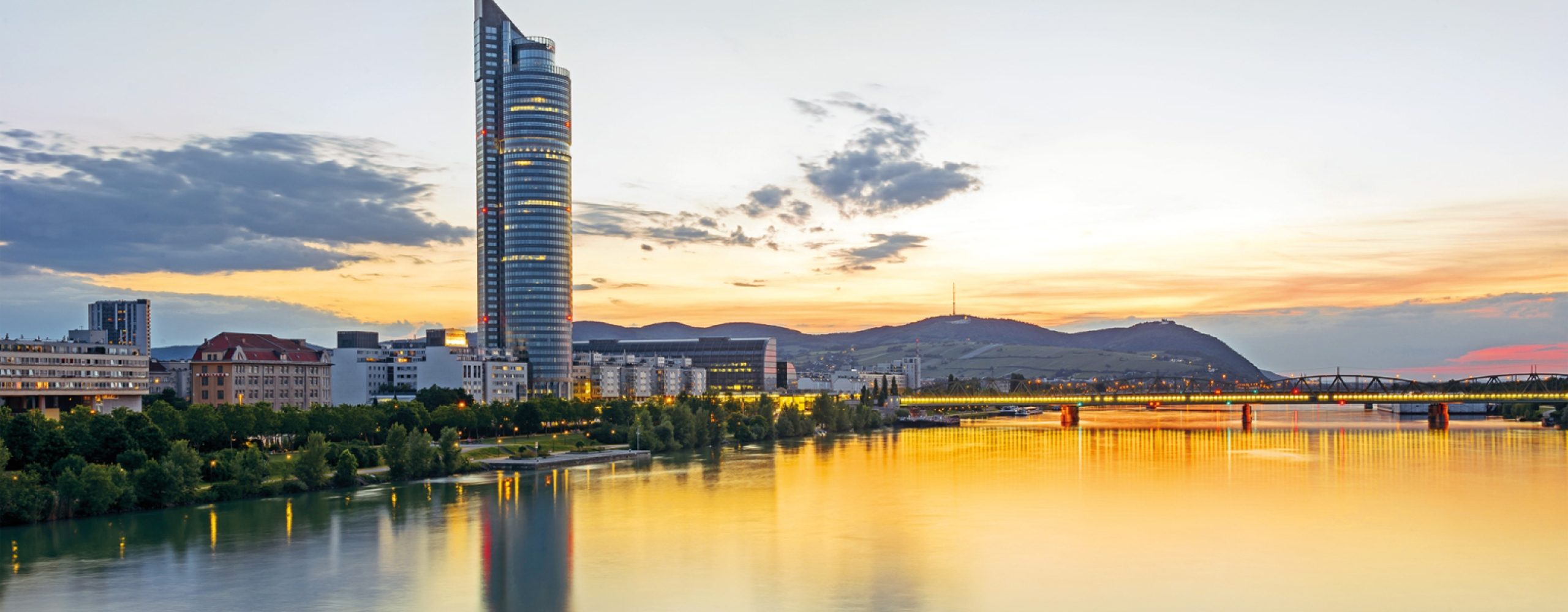 Der Millennium Tower Wolkenkratzer in Wien, Österreich, mit Blick auf die Donau während eines goldenen Sonnenuntergangs und Bergsilhouetten im Hintergrund.