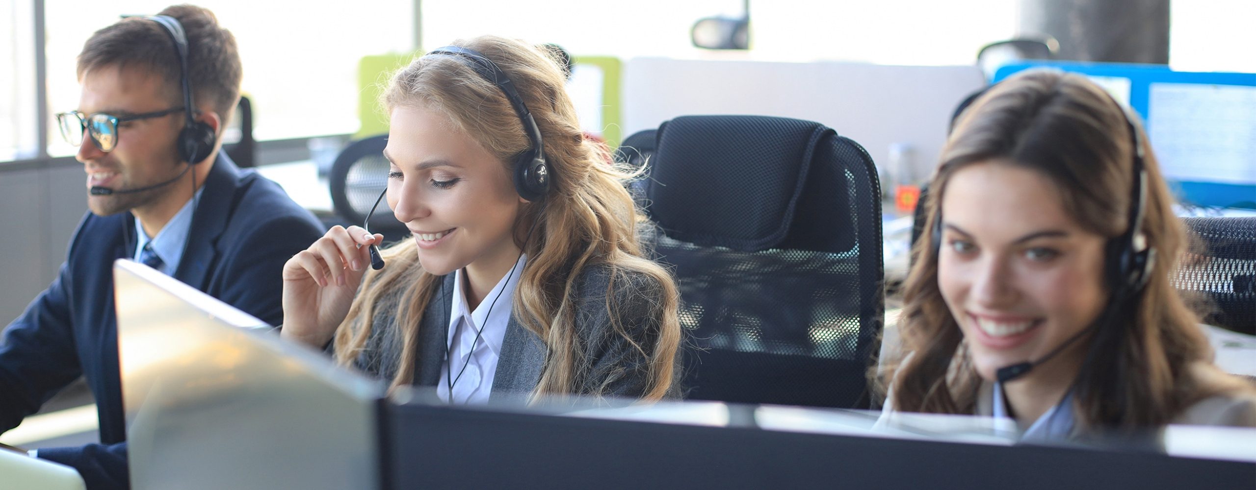 Drei lächelnde Kundenservice-Mitarbeiter mit Headsets arbeiten an ihren Computern in einem hellen Büro.